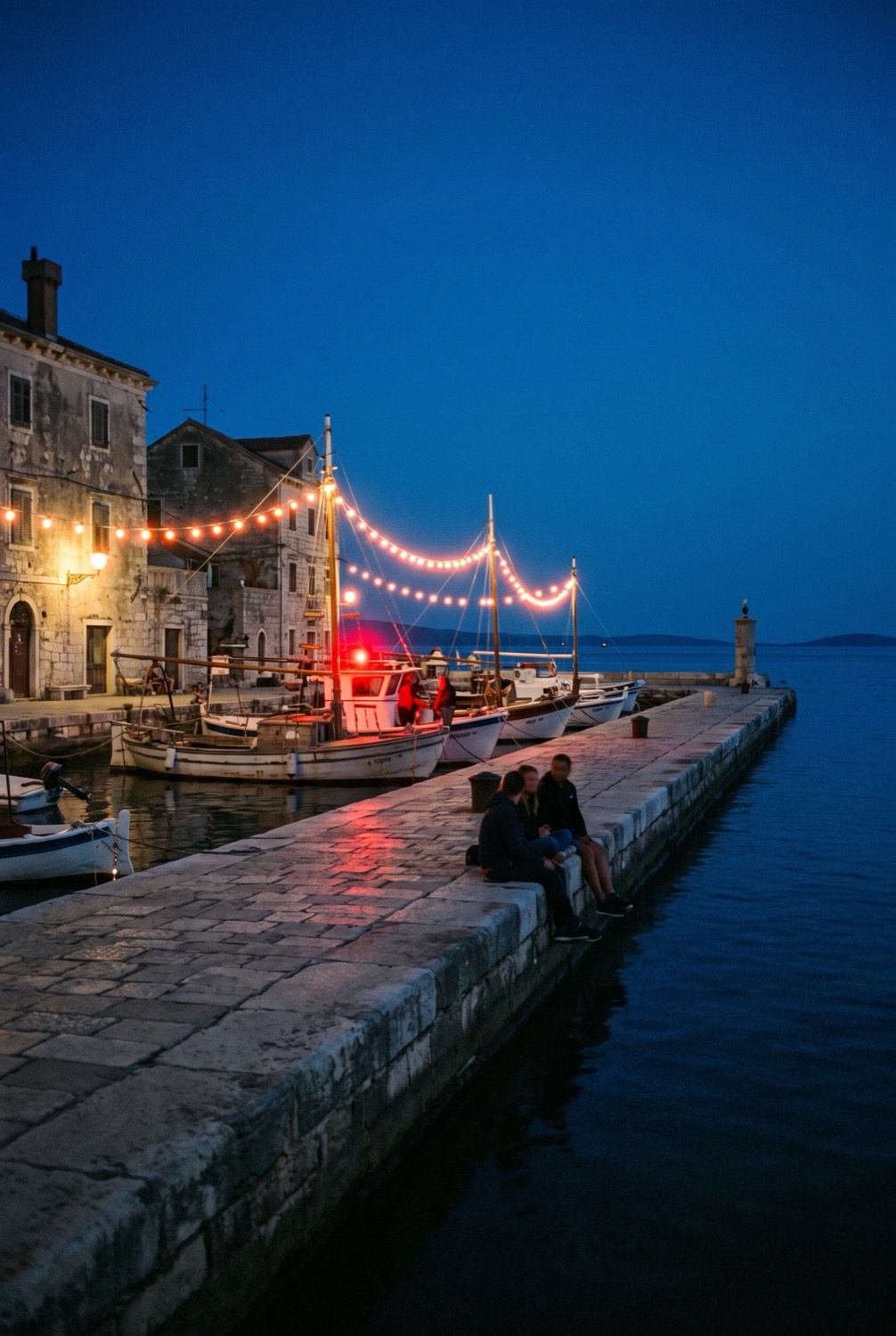 A stone pier running out into the sea at dusk, string lights over moored boats