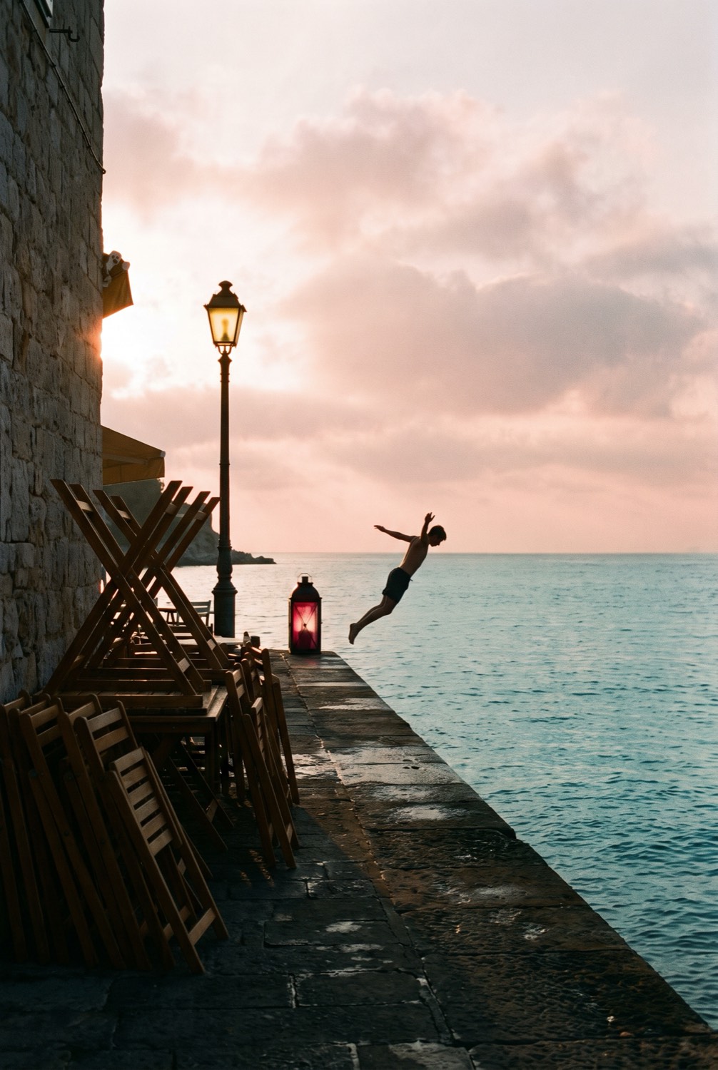 A swimmer diving off the end of the pier at sunrise, chairs stacked on the quay