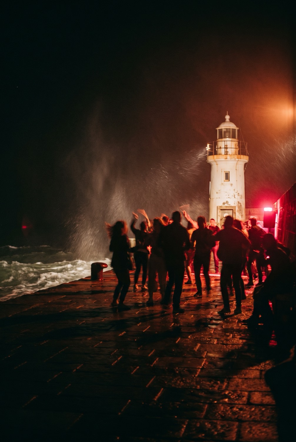People dancing by the lighthouse in red light, spray coming over the sea wall