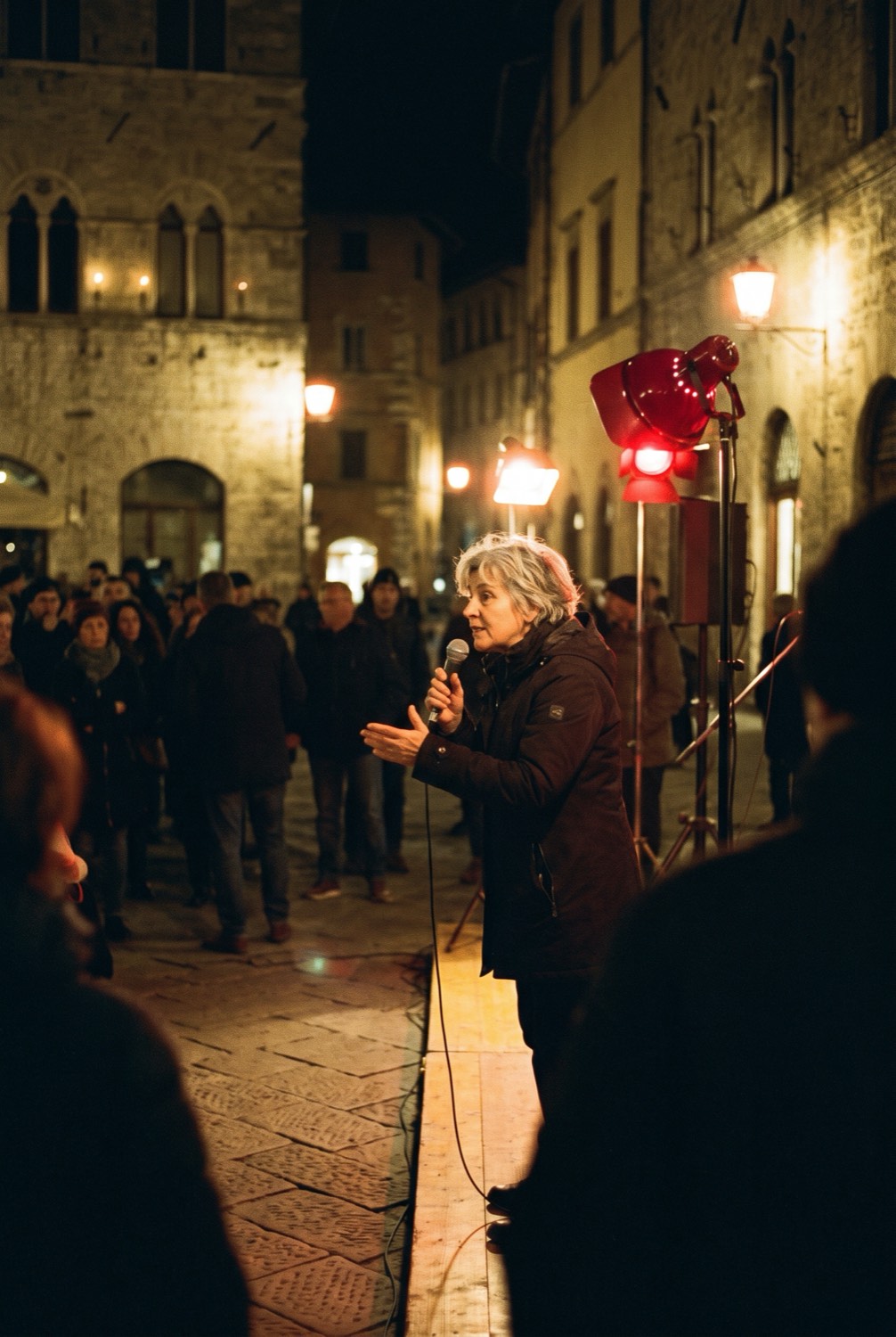 A woman speaking into a microphone on a low stage in a stone square at night